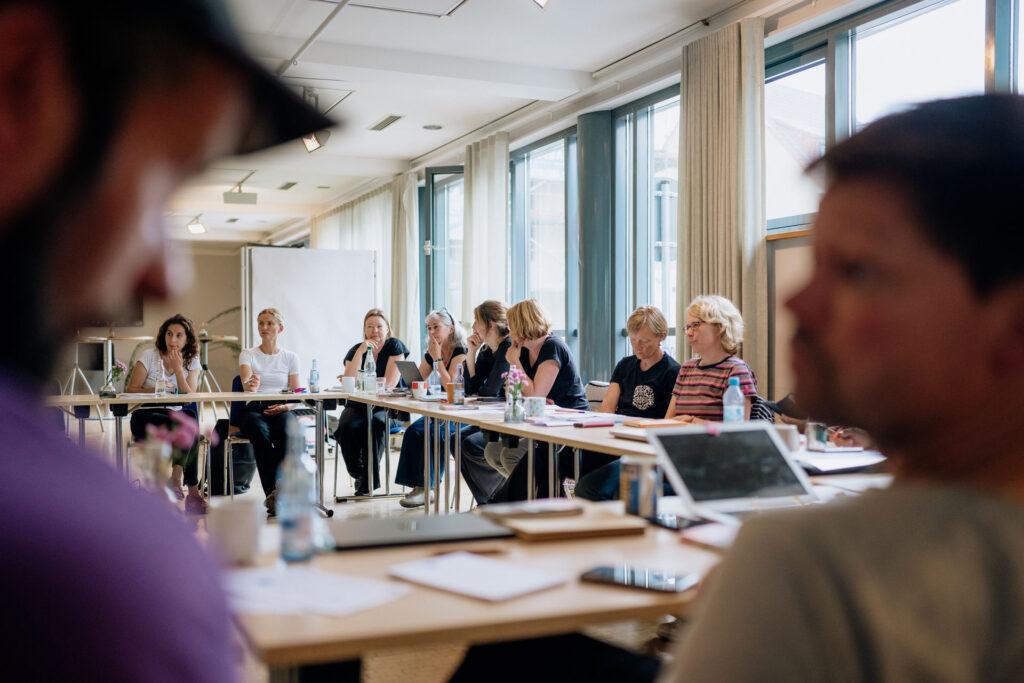 Mehrere Personen sitzen an zusammengestellten Tischen in einem hellen Seminarraum und nehmen einer Besprechung teil. Auf den Tischen stehen Laptops, Notizbücher und Getränke. Im Vordergrund unscharf ein Mann, im Hintergrund fokussiert eine Gruppe konzentrierter Teilnehmer:innen.