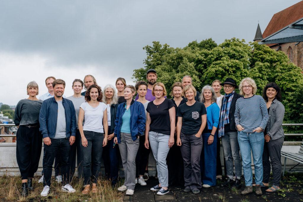Gruppenfoto von etwa 20 Personen auf einer begrünten Dachterrasse oder Außenfläche mit Bäumen und einem historischen Gebäude im Hintergrund. Die Personen stehen eng beieinande und, lächeln in die Kamera. Sie sind Vertreter:innen der KUlturverbände und Fachstellen in Mecklenburg-Vorpommern